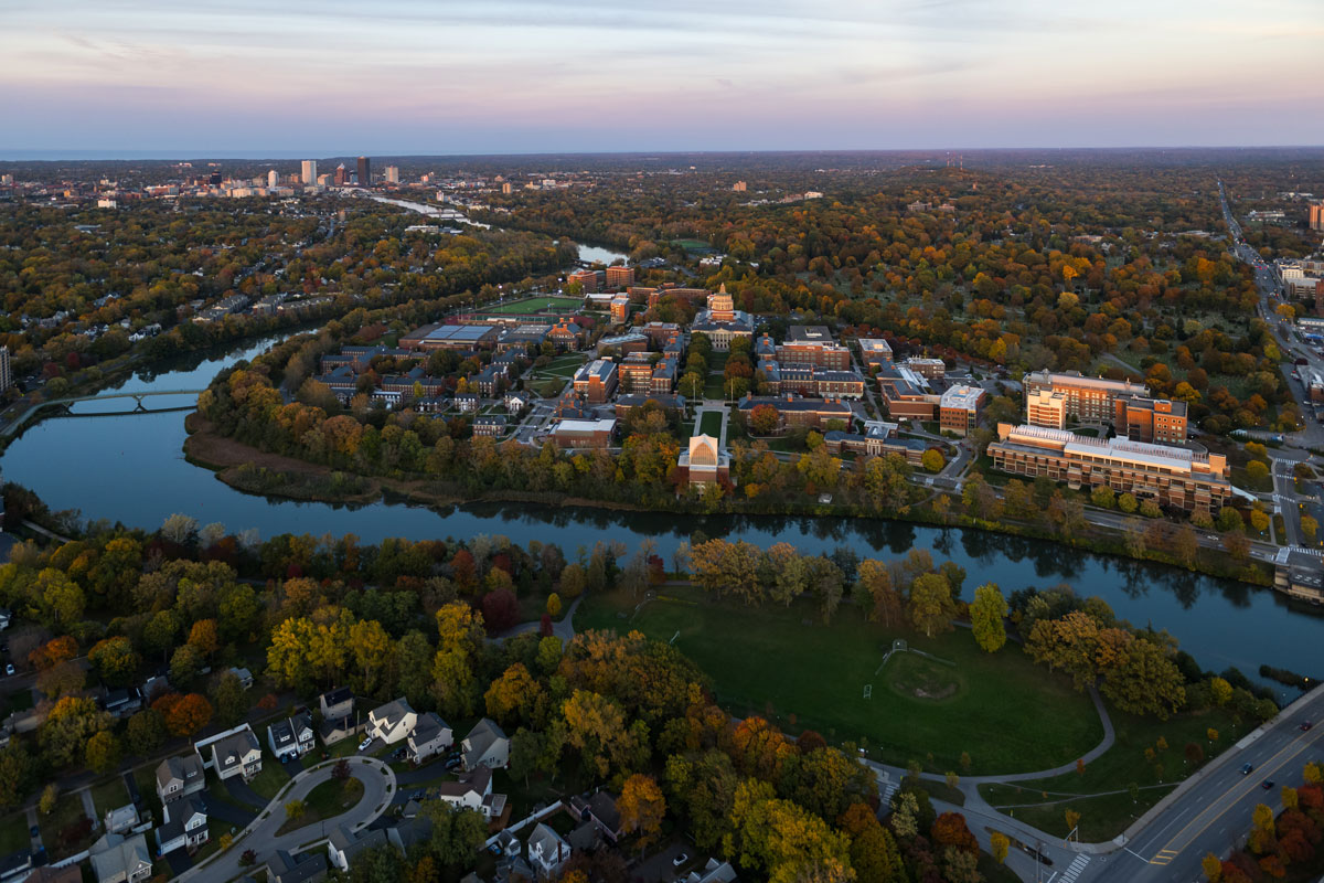 罗彻斯特大学广阔的校园鸟瞰图，前景是一条蜿蜒的河流, 周围是秋天的树木. 对岸以郊区社区为特色, 在多云的天空下，可以看到远处城市建筑的天际线.
