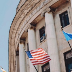关闭-up view of a neoclassical government building with large columns and a facade made of stone. 几个旗帜, 包括美国国旗, are visible in front of the building against a clear blue sky.