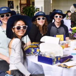 A group of smiling young adults sits at a table, each wearing white sunglasses and matching blue hats. They are surrounded by gift boxes, pamphlets, and floral arrangements. The setting appears to be a casual event or gathering.