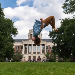 A person is mid-air performing a backflip on a grassy field with a large, stately building with columns and a dome in the background. The sky is partly cloudy and there are several people and trees in the vicinity.